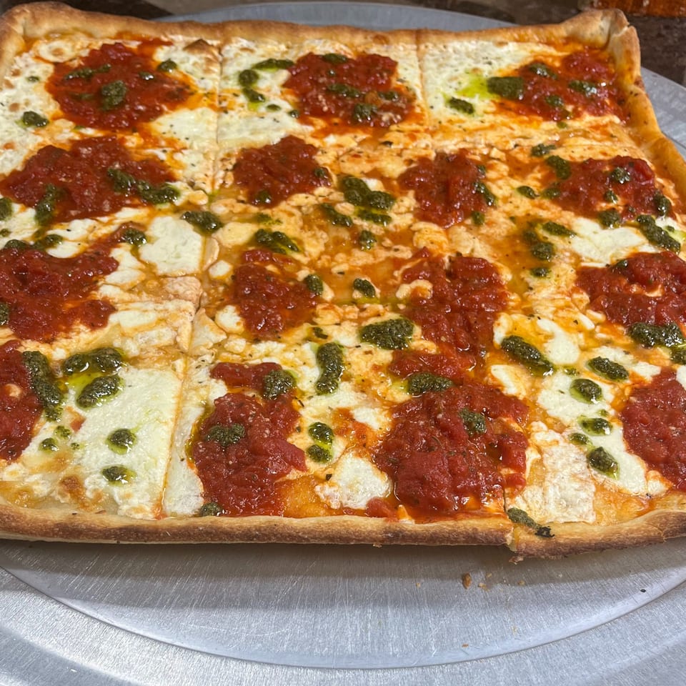 A rectangular Sofia Pizza pie with red sauce, white mozzarella, and green pesto on a metal tray.