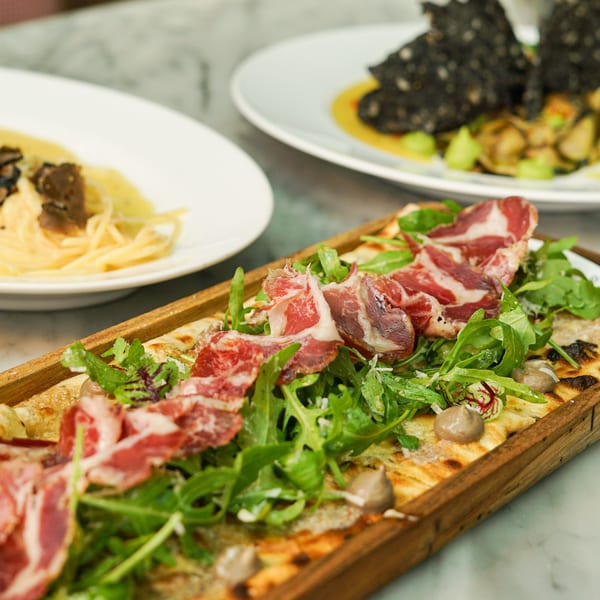 Italian flatbread with prosciutto, arugula, and sauce, with pasta and another dish in the background.