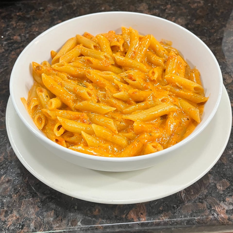 A white bowl filled with penne pasta in a creamy orange sauce, served on a white plate.