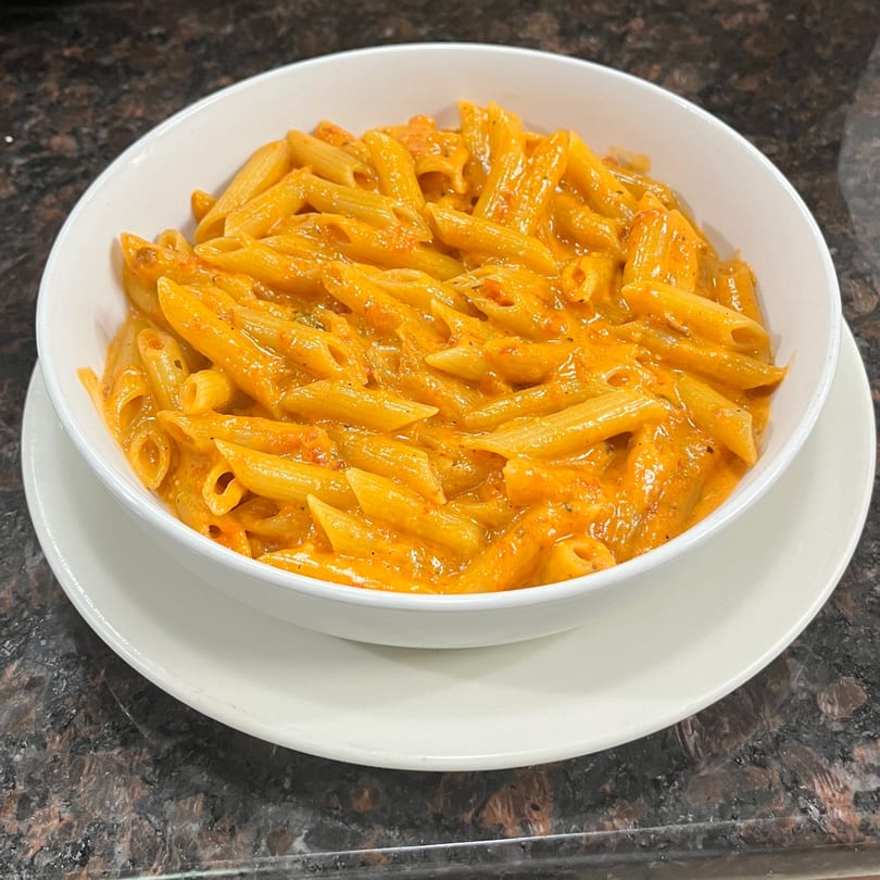 A white bowl filled with penne pasta in a creamy orange sauce, served on a white plate.