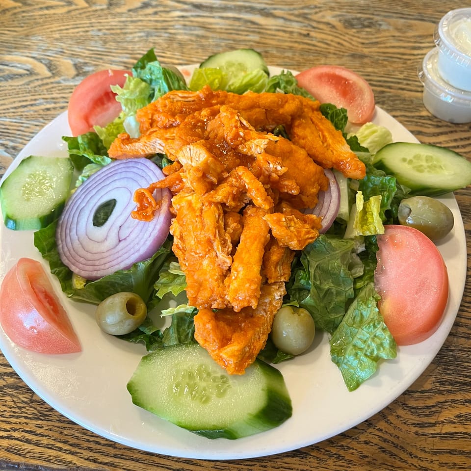 Buffalo chicken salad with lettuce, tomatoes, cucumbers, red onions, and olives on a white plate.